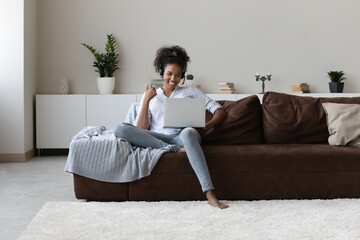 Smiling African American woman in headset using laptop, sitting on couch at home, young female...