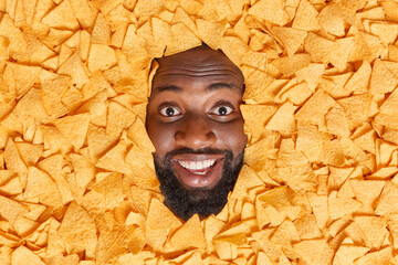 Cheerful dark skinned man with thick beard buried in delicious mexican nachos chips enjoys eating tasty salty spicy snack smiles broadly. Positive guy surrounded by triangular crunchy crisps - obrazy, fototapety, plakaty
