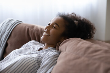 Close up excited African American woman dreaming with closed eyes, leaning back on cozy couch, smiling young female enjoying lazy leisure time weekend at home, daydreaming and visualizing future