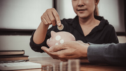 Men and women put dollar coins in a piggy bank and write household income and expenditure records,...