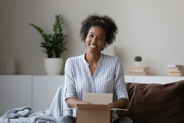 Portrait smiling African American millennial woman unpacking parcel box with order on couch at home, satisfied young female customer buyer excited by good quick delivery service, shipping concept