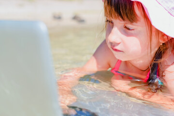 Rest and productive distance Online studying. Humorous portrait of little cute girl learning remotely with laptop outdoors in tropical sea. Horizontal image.