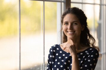 Portrait of happy millennial Latin 30s woman touching chin, facial skin with toothy smile, standing by window, looking at camera. Head shot of beautiful young Hispanic female model in home interior