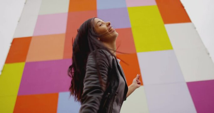4K Beautiful , stylish , young girl with black hair  beautiful body in Leather Jacket and sport jeans  dancing , moving , smiling and looking at camera with happy emotions . Slow motion colorful wall