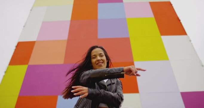4K Beautiful , stylish , young girl with black hair  beautiful body in Leather Jacket and sport jeans  dancing , moving , smiling and looking at camera with happy emotions . Slow motion colorful wall