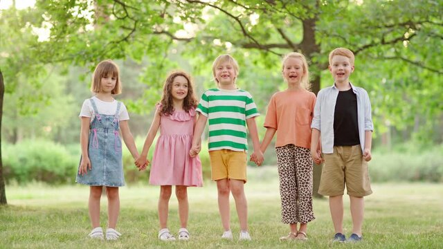 Zoom in of five cute kids standing holding hands on grass in green park, looking at camera and screaming happily. Portrait of excited little friends having fun together