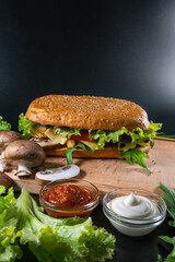 Fresh and tasty Italian panini, sandwich with ham and vegetables, sauce, salad, mushrooms, tomato and cucumber, on wooden spacing, plate. Black background. Vertical photo. Copy space