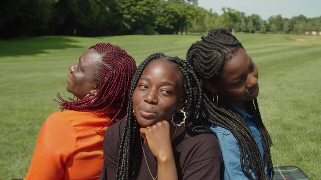 Portrait of joyful happy pretty african mother and three adorable teenage daughters with braided hair sitting back to back in circle on green lawn, enjoying leisure together and communicating in park.