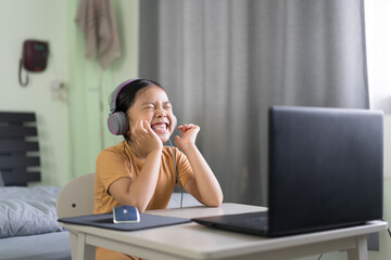 Asian child or kid girl smile enjoy e-learning on computer notebook with wearing headphone for laugh to funny communication and study online or people learn from home to play laptop by back to school