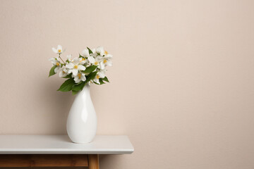Fototapeta na wymiar Bouquet of beautiful jasmine flowers in vase on table near beige wall, space for text