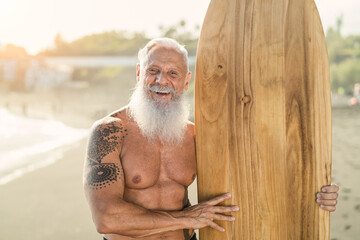 Senior man having fun surfing during sunset time - Fit retired male training with surfboard on the beach - Elderly healthy people lifestyle and extreme sport concept