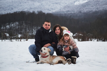 Fototapeta na wymiar Vacation with family during New Years Christmas holidays. Young cute Caucasian family mom dad and daughter with white fluffy half breed shepherd dog are sitting in winter park in snow and smiling.
