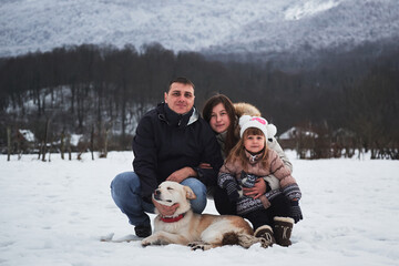 Vacation with family during New Years Christmas holidays. Young cute Caucasian family mom dad and daughter with white fluffy half breed shepherd dog are sitting in winter park in snow and smiling.