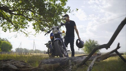 Brutal young man in sunglasses, blue sits on a custom motorcycle on the field with a black helmet. Motorcyclist on the meadow in the summer. Motorcyclist sitting near a motorcycle