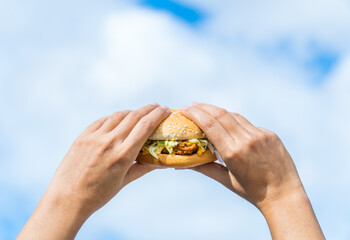 close up of tasty burger. hand holding juicy hamburger on sky background. concept hamburger at street food festival. summertime. summer vacation picnic. space for text
