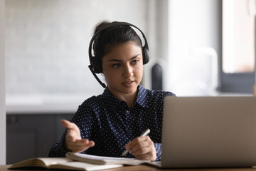 Indian female student in headphones watching and listening webinar from home, writing notes,...