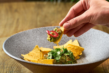 Mexican avocado guacamole with nacho, hot pepper and basil in a gray plate. hand dips nacho in guacamole