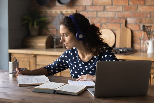 Focused student woman using headphones and laptop, studying from home, reading book, doing homework research, listening webinar, learning seminar, tutor guidance, writing notes