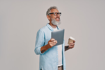 Happy senior man holding digital tablet and coffee cup while standing against gray background