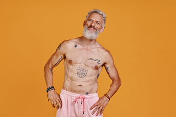 Shirtless tattooed senior man looking away and smiling while standing against orange background