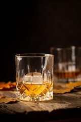 Glass of scotch whiskey and ice on wooden background with autumn leaves