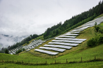 Solar, photovoltaic power station located on a mountain slope in the Alps. Foggy day, green pasture grass and blue skies and mountains create a fresh and ecological image. - obrazy, fototapety, plakaty