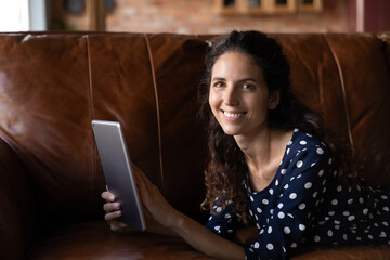 Happy woman relaxing on couch with tablet, holding device, looking at camera, smiling, watching video, movie, reading book. Student using digital pad for learning, attending virtual class from home
