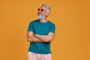 Stylish senior man keeping arms crossed and smiling while standing against orange background