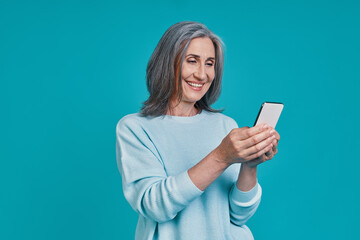 Fototapeta premium Mature beautiful woman using smart phone and smiling while standing against blue background