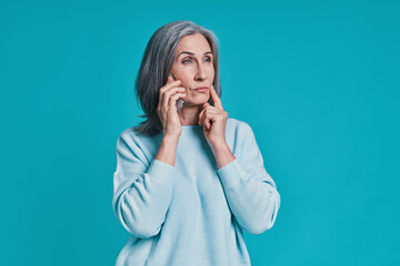 Mature beautiful woman talking on smart phone while standing against blue background