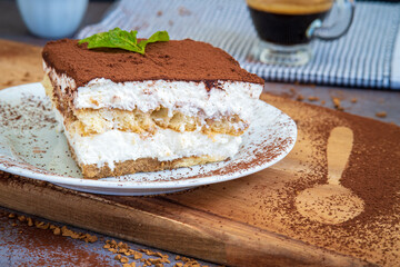 Slice of traditional tiramisu cake served with a cup of coffee in the background. Authentic gluten-free Italian layered dessert with ladyfinger biscuits, mascarpone cheese cream and cocoa powder. Side
