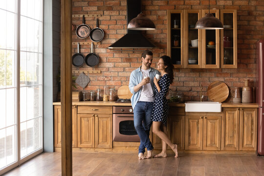 Happy young married couple enjoying morning coffee in kitchen of new apartment. Barefooted dating guy and girl standing together on heat floor at home, hugging, drinking hot beverage. Full length