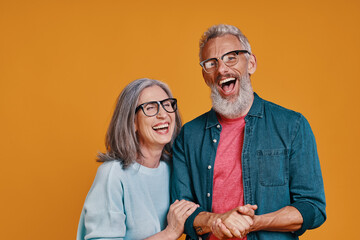 Happy senior couple in eyeglasses smiling while standing together against orange background