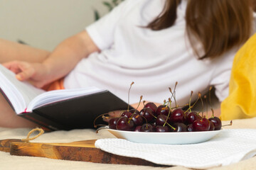A young woman is lying with a book on the bed near a plate of cherries.