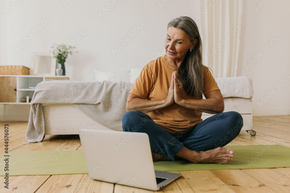 Wall mural Calm middle-aged woman holding hands in namaste gesture. Mature yoga teacher practicing meditation online at home. Zen, balance and self-awareness concept - Wall murals