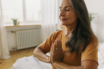 Calm middle-aged woman sitting in padmasana with eyes closed. Mature female holding hands pressed...