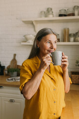 Smiling middle-aged woman drinking tea in home kitchen. Cheerful elderly female enjoying morning...