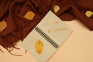 Atmospheric autumn composition. Fall background with notepad with Hello Autumn text, deep brown cashmere scarf and leaves on pastel colors. Sunday relaxing, memories and still life concept. Flat lay