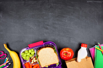 Healthy school lunch box and school supplies. Bottom border, top view on a chalkboard background.