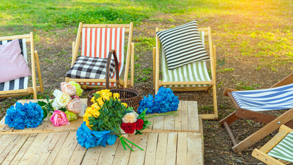 Beautiful artificial bouquets of various colors placed on wooden table among deck chairs in garden. Multi-colored artificial flowers adorn the wooden table for beautiful and relaxing. Selective focus.