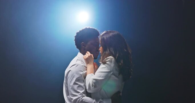 Romantic shot of couple dancing, expressing love, beautiful woman touches dark skinned handsome man on neck, guy tries to kiss her but moves away after a while, background lit up with blue flash