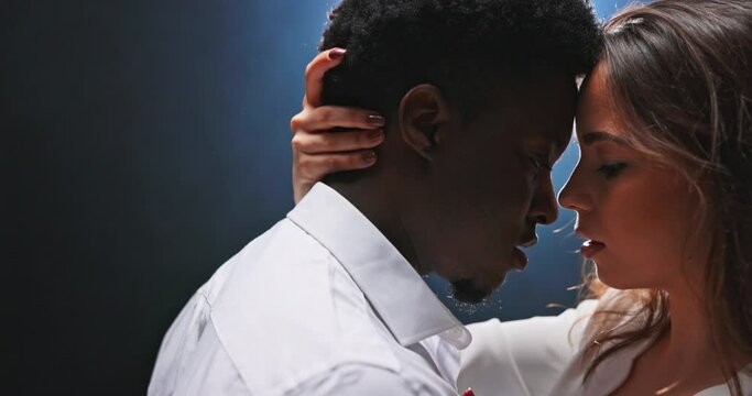 Close-up of couple's faces looking deep into each other's eyes with solemnity while dancing together, dark-skinned men charms woman who gently runs hand over partner's neck. Flare on dark background