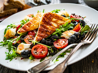 Fresh vegetable salad with grilled halloumi cheese, tomatoes,  lettuce, green leaves, black olives and onion on wooden table
