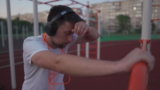 Fit man with headphones after physical training jumping rope at city stadium. Male taking break after exercising session listening music on headphones with skipping rope around neck at outdoor gym.