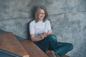 Senior beautiful woman using smart phone and smiling while sitting on the stairs indoors