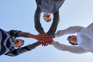 A low angle shot of three young friends putting their hands together against a blue sky