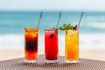 Set of Three Refreshing Fruit Cocktails Standing on Table on Beach, Blurred Turquoise Sea on Background. Colorful Summer Ice Cold Drinks. Fresh Healthy Beverages with Berries, Orange and Passion Fruit