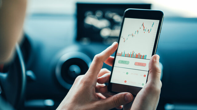 Woman Using White Mobile Phone Investing Application In Car. Stock Market Investment App In Hand On The Go. Close-up View