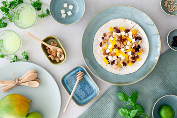 Flat lay of a delicious tortilla wrap with feta cheese, yellow paprika, raisins and cashew nuts with honey, surrounded by blue-green ceramic plates and bowls with a mango in the left hand corner.