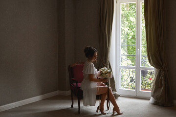 A young beautiful bride in silk peignoir holds wedding bouquet and sits on the chair in a hotel room